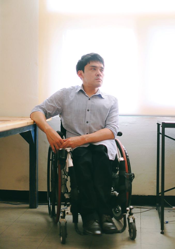 ウォニョンさんの写真。手動車椅子に乗り、 ブラインダー越しに光が差しこむ窓の前にすわり、机に片肘を置いた状態でななめ前を見つめている。